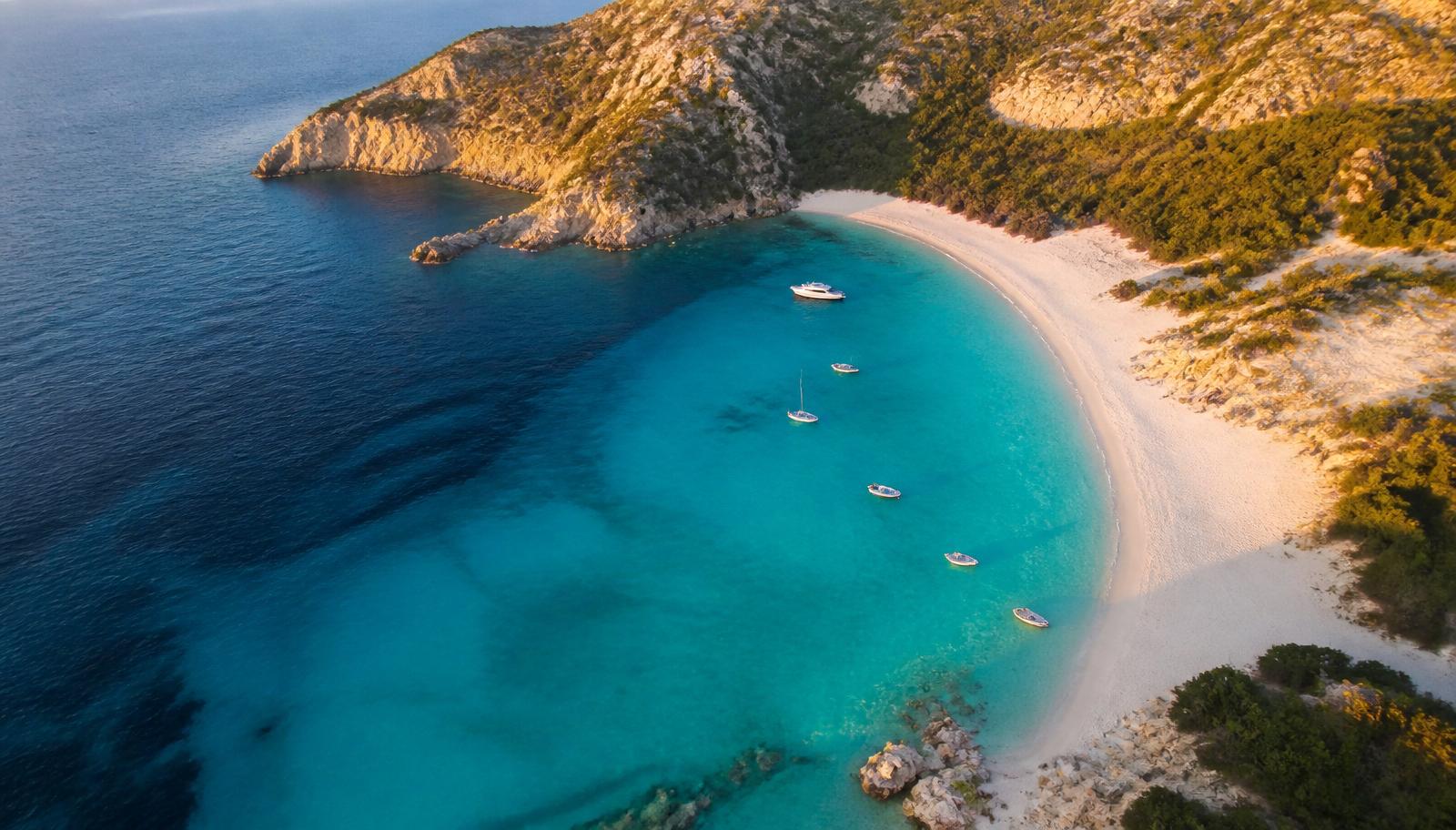Aerial view of a Mediterranean crescent beach with turquoise water and small boats at golden hour